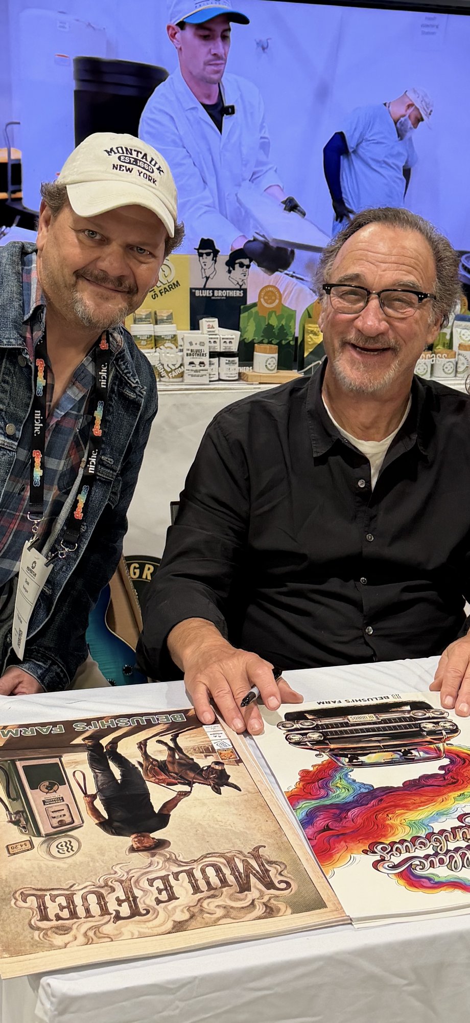 Jim Belushi signing Belushi's Farm posters (Mule Fuel / Cadillac Rainbows) on the NECANN floor — TSR press badge visible